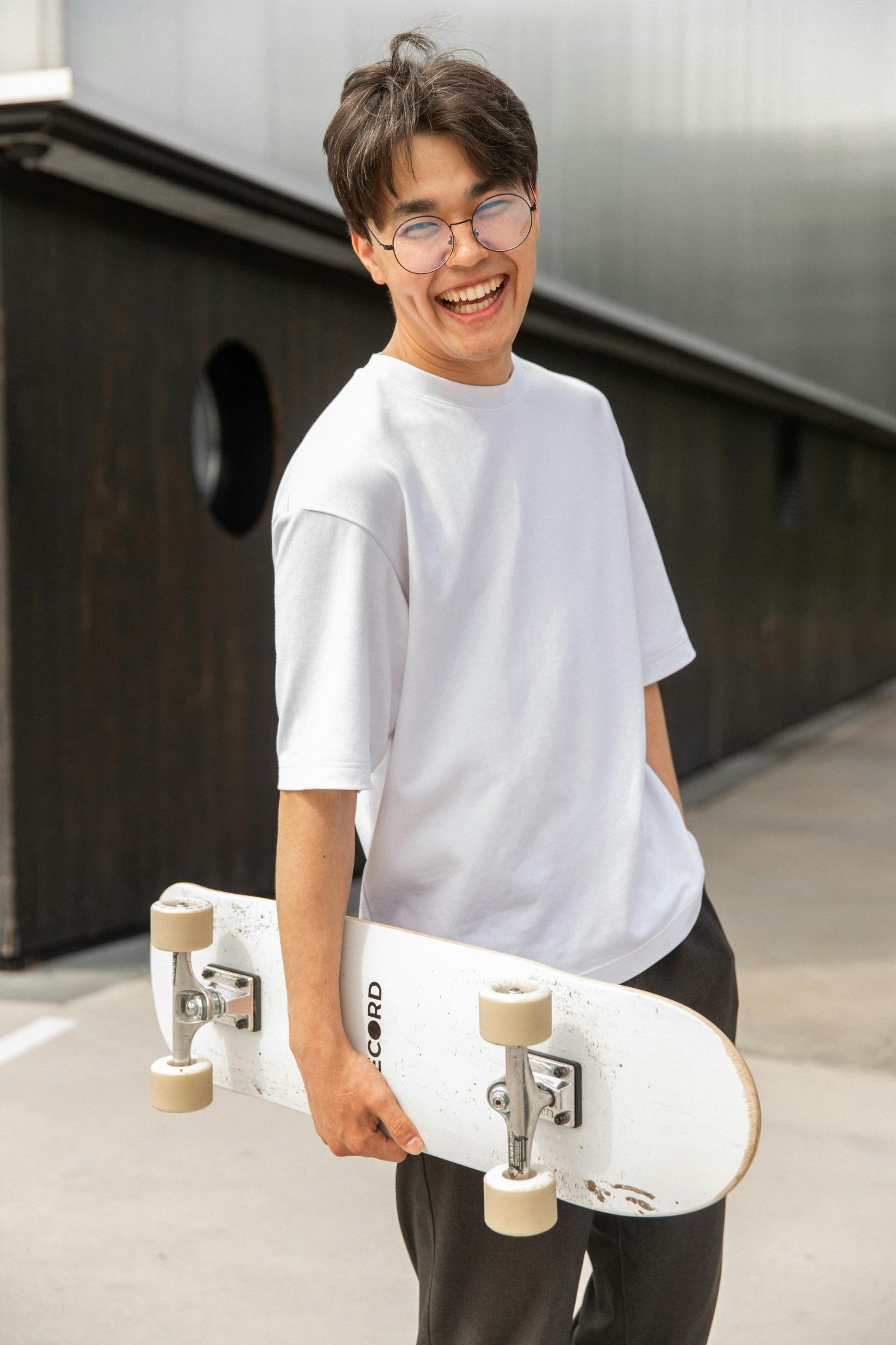 Happy young man with glasses holding a skateboard at a skatepark, laughing with joy.